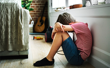 A boy sits sadly on the floor in his home, back against the wall, head on his knees